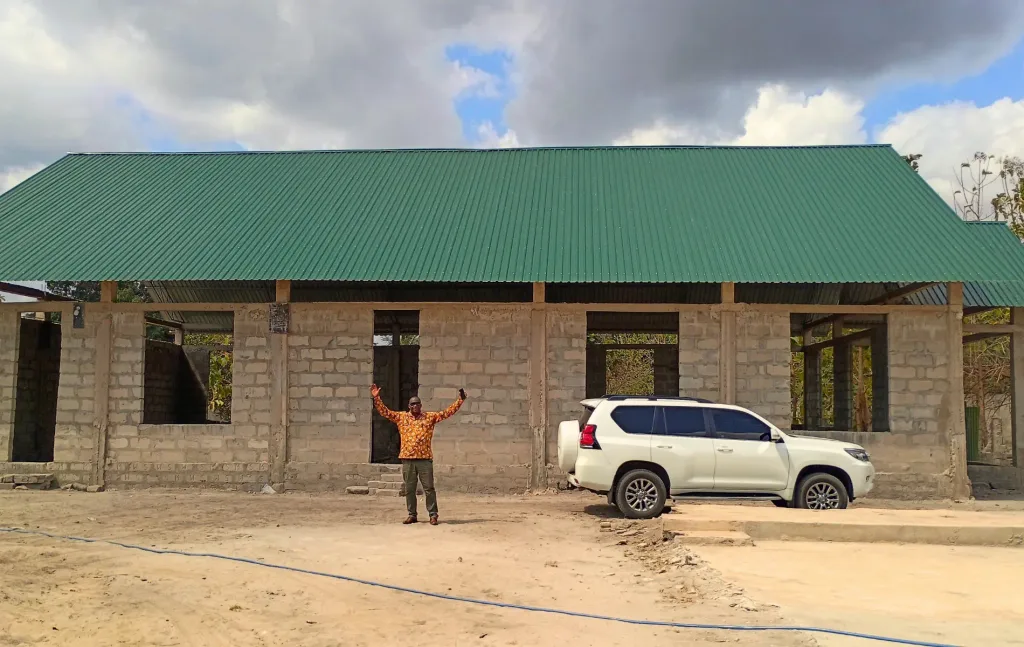 Emmanuel stands with his arms raised outside the unfinished Kiromo Church in Tanzania. The large building has a green metal roof and open window and doorway spaces.