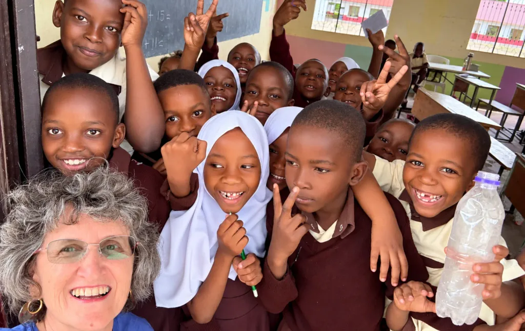 Heidi smiles alongside a lively group of Lawrence Academy students in Tanzania. Children crowd together inside a classroom, laughing, smiling, and making playful gestures toward the camera.