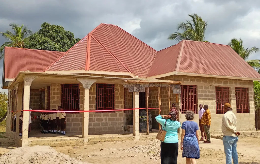 The unfinished Kiromo Medical Clinic in Tanzania during its dedication ceremony. Students gather beneath the covered entrance while AFAC representatives and local partners stand outside the building.