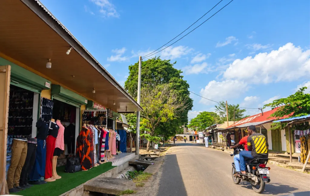 A motorcycle carrying two riders travels along a paved street lined with clothing shops and market stalls in Tanzania. Buildings, trees, and overhead power lines stretch into the distance beneath a blue sky.